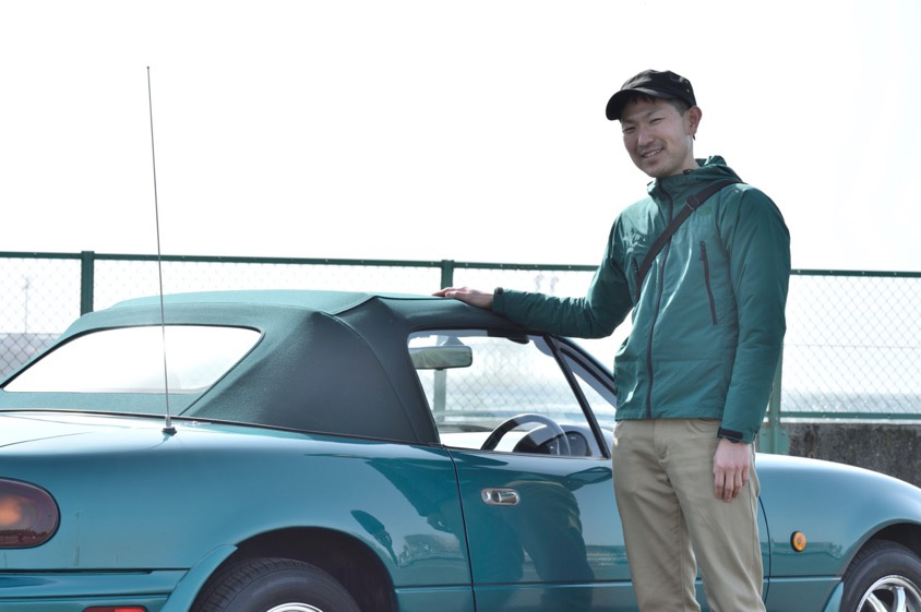マツダ・ロードスター（NA型）の横でほほ笑むオーナーの秋山さん