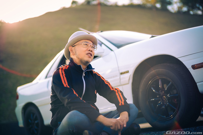 いすゞ・ピアッツァの前に座って話す横田をさむさん